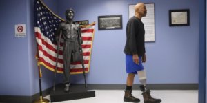 A veteran with a prosthetic leg walks past a statue with only on leg in front of an American flag