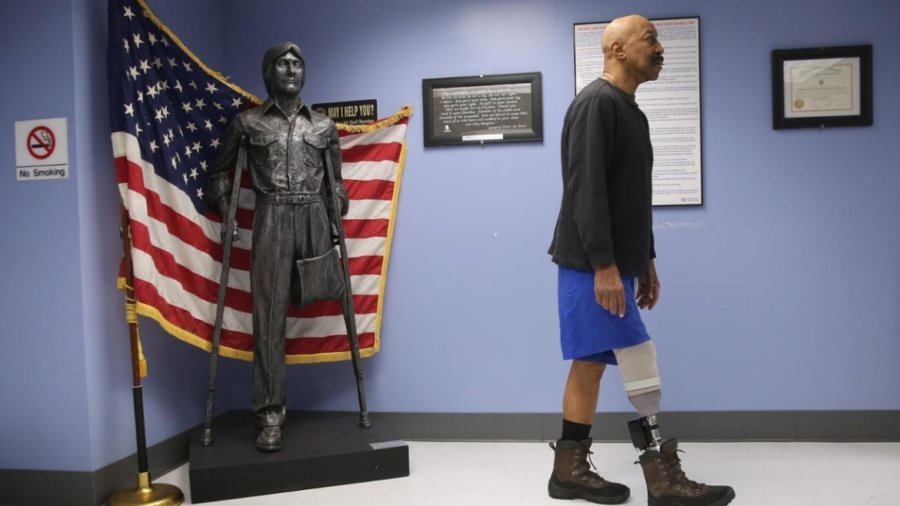 A veteran with a prosthetic leg walks past a statue with only on leg in front of an American flag