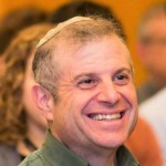 headshot of David B. Marcu smiling wearing a green collard shirt with people behind him