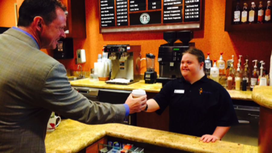 a woman with a disability working as a barista offers a customer a coffee as an example of the #RespectTheAbility campaign