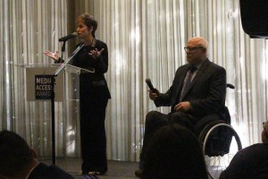 Deborah Calla standing behind podium and Allen Rucker seated in wheelchair on stage