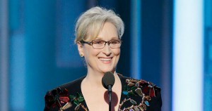 Meryl Streep standing behind a microphone smiling. She is wearing a black dress with many colorful, light-reflecting jewels.