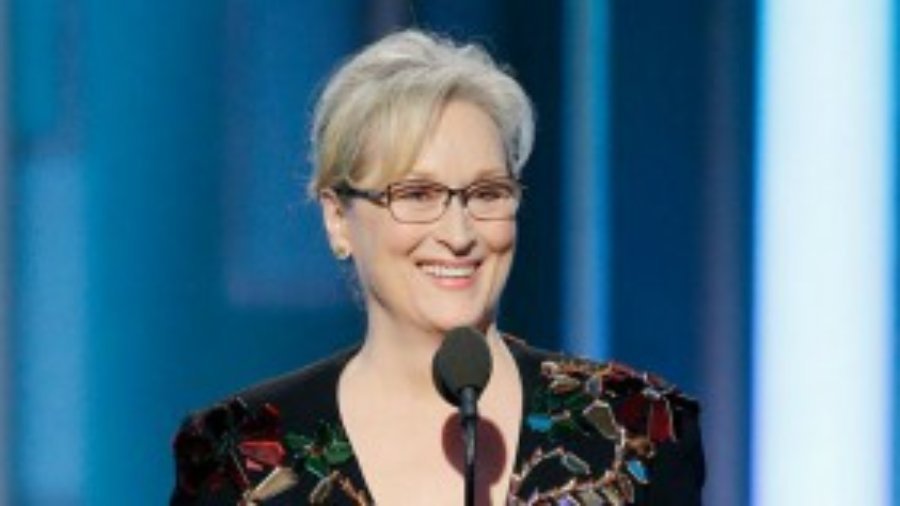 Meryl Streep standing behind a microphone smiling. She is wearing a black dress with many colorful, light-reflecting jewels.