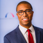 head shot of Calvin Harris wearing a blue suit, white shirt and red tie