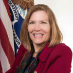Jennifer Sheehy official DOL Portrait wearing a red blazer sitting in front of two flags - American flag and DOL flag
