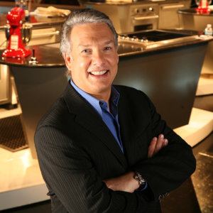 headshot of Marc Summers wearing a black suit and blue shirt with arms crossed in front of a kitchen set