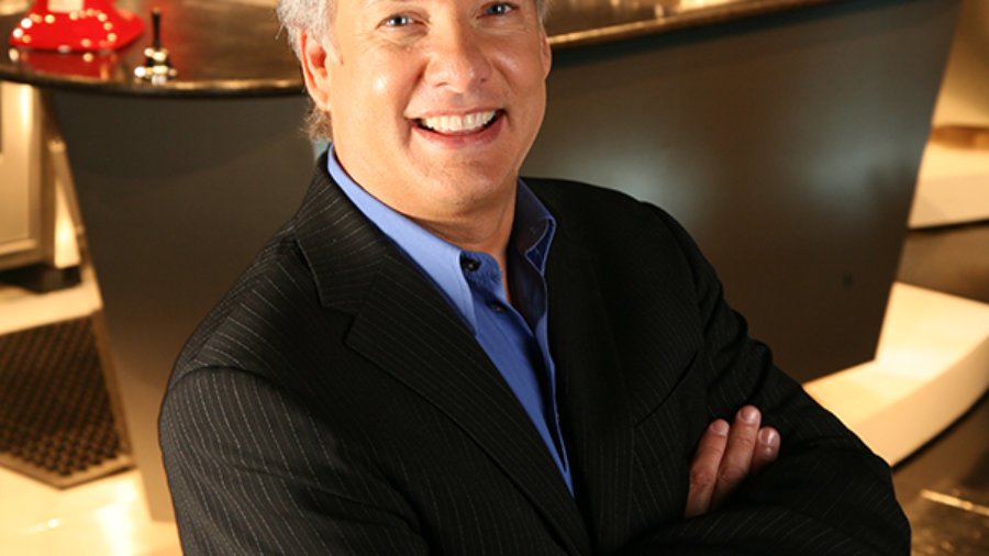 headshot of Marc Summers wearing a black suit and blue shirt with arms crossed in front of a kitchen set