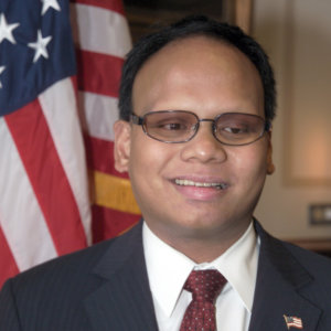 Ollie Cantos smiling in an official portrait wearing a suit and American flag pin in front of an American flag
