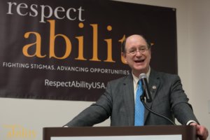 Brad Sherman speaking at the microphone with a RespectAbility banner behind him