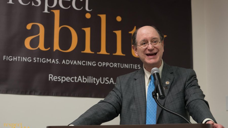 Brad Sherman speaking at the microphone with a RespectAbility banner behind him