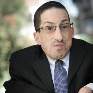 headshot of Steven James Tingus wearing a black suit and blue tie seated in his wheelchair outside color photo