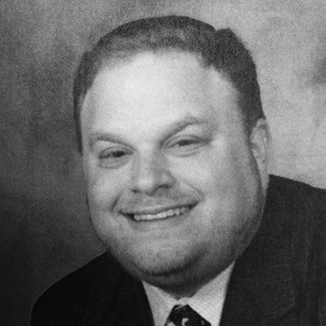 headshot of Aaron Orlofsky smiling and squiting eyes, wearing suit and tie grayscale photo