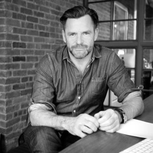 Andrew Egan leaning on a table wearing a button down shirt and facing the camera grayscale photo