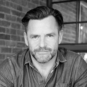 headshot of Andrew Egan leaning on a table wearing a button down shirt and facing the camera grayscale photo