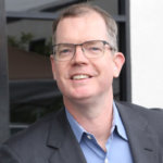 Delbert Whetter smiling wearing a suit jacket and glasses