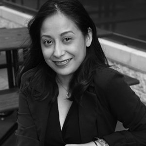 Marisela is sitting on bench smiling with a black dress and black blazer on grayscale photo