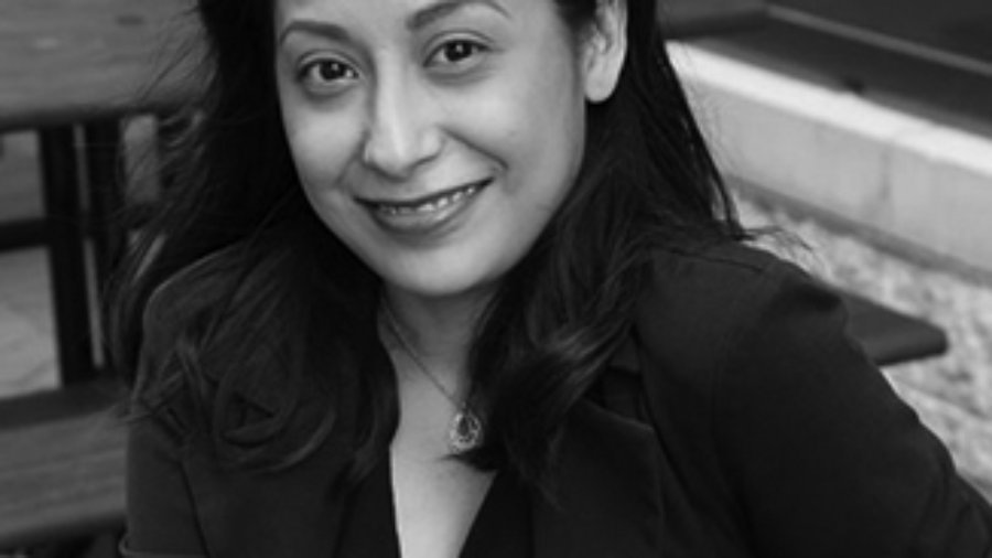 Marisela is sitting on bench smiling with a black dress and black blazer on grayscale photo