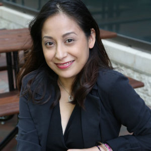 Marisela is sitting on bench smiling with a black dress and black blazer on