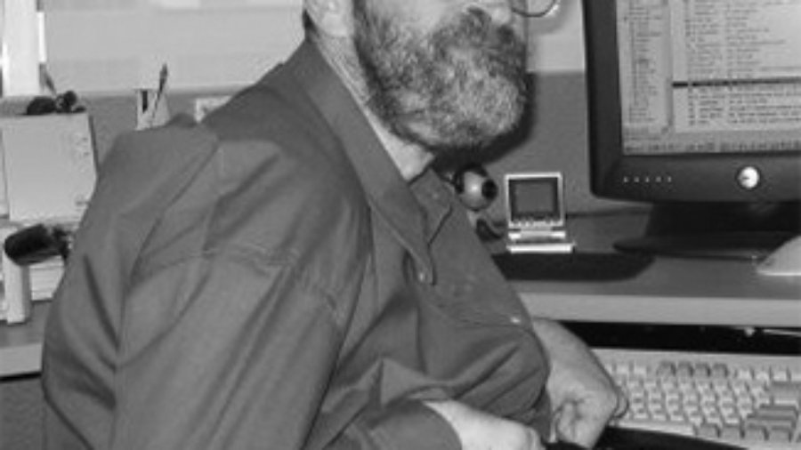 Neil Jacobson sitting at his computer and smiling he has a beard and is wearing glasses grayscale photo