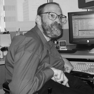 Neil Jacobson sitting at his computer and smiling he has a beard and is wearing glasses grayscale photo