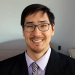 Randall Duchesneau smiling and wearing a black suit, blue shirt, and black and white checkered tie and glasses color photo