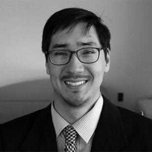 Randall Duchesneau smiling and wearing a black suit, light shirt, and checkered tie and glasses grayscale photo