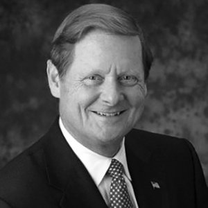 Steve Bartlett is smiling and wearing a black suit, white shirt, a spotted tie and an american flag pin on his jacket, grayscale photo