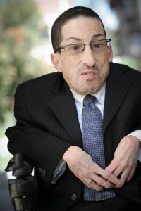 Steven James Tingus wearing a black suit and blue tie seated in his wheelchair outside