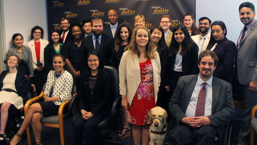 The whole RespectAbility team, so the staff, and all of the fellows are posed in front of the wall that has the RespectAbility logo printed on it. The photo is in color.