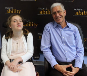 Lilly Grossman seated in her wheelchair and Richard Wolf seated in a chair smiling in front of a RespectAbility banner