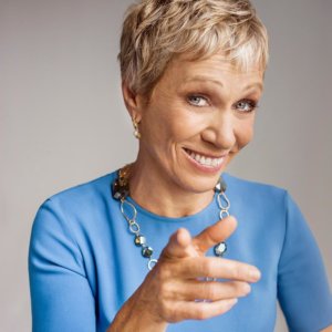 Barbara Corcoran pointing toward the camera wearing a blue top and silver necklace