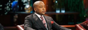 Daymond John in a black suit seated on the set of Shark Tank