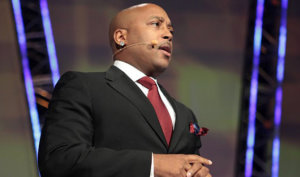 Daymond John in a black suit and red tie speaking into a microphone