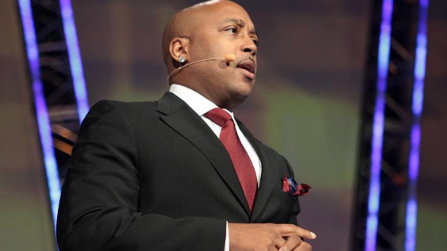 Daymond John in a black suit and red tie speaking into a microphone