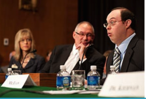 Egan seated behind a table with other panelists, with bottles of water and nameplates in front