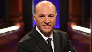 Kevin O'Leary posing for the camera wearing a black suit on the set of Shark Tank