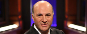 Kevin O'Leary posing for the camera wearing a black suit on the set of Shark Tank