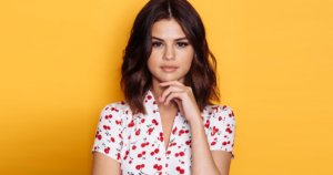 Selena Gomez wearing a white blouse with a cherry print posing for the camera with a yellow background
