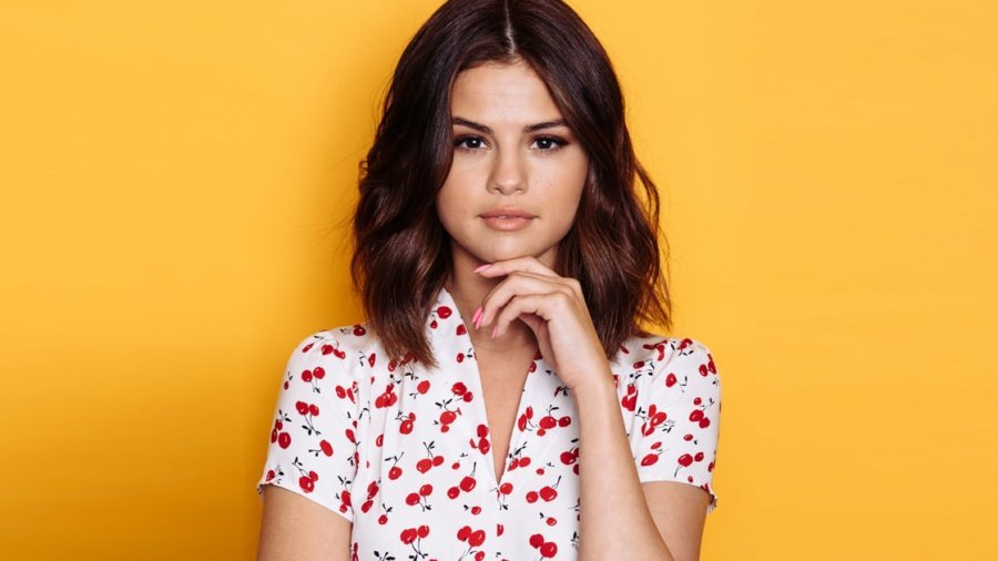 Selena Gomez wearing a white blouse with a cherry print posing for the camera with a yellow background