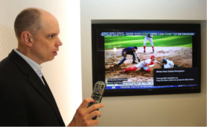 Tom Wlodkowski holding a remote in front of a wall mounted TV showing a baseball game