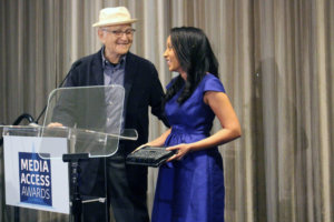 Norman Lear with his hand on host Haben Girma's shoulder, speaking behind the podium