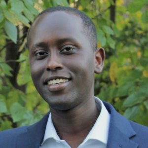 Ian Cherutich smiling wearing a navy blue suit in front of trees and bushes outside