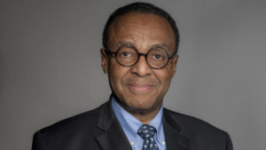 Clarence Page headshot wearing black suit, blue shirt and glasses