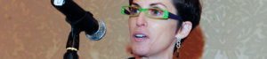 close up of Deborah Calla's face wearing green glasses behind a microphone