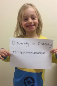 Maisie Sly smiling and holding a sign that reads "Disability is Diversity #thesilentchildoscars90"