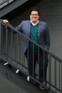 A Latino man standing on steps posing for the camera wearing a gray suit and green sweater