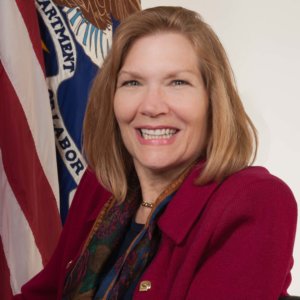 Jennifer Sheehy headshot, smiling in front of an American flag and a Department of Labor flag