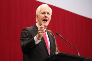 Sen. John Cornyn giving a speech at a podium in front of a red and white background