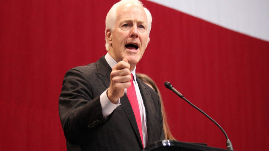 Sen. John Cornyn giving a speech at a podium in front of a red and white background