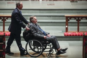 President George H.W. Bush wearing John's Crazy Socks to Barbara Bush's funeral. Bush is in a wheelchair being pushed by his son, President George W. Bush.